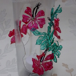 Könnte beinhalten: Eine klare Glasvase mit strukturierter Oberfläche ist mit handbemalten rosa Hibiskusblüten und grünen Blättern verziert.