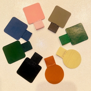 Może przedstawiać: Zestaw ośmiu kolorowych skórzanych uchwytów do stron notesu. Uchwyty mają kształt kwadratów i kół. Kolory obejmują różowy, zielony, niebieski, brązowy, beżowy i pomarańczowy.