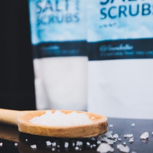 May include: A wooden spoon filled with white salt crystals, with two bags of salt scrubs in the background. The bags are labelled "Salt Scrubs".
