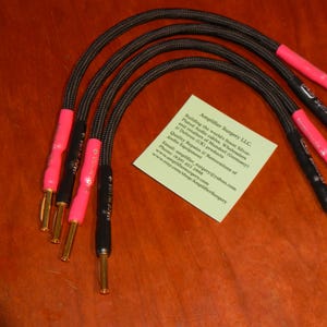 May include: Four black speaker cables with pink insulation and gold-plated connectors. The cables are coiled and arranged in a semi-circle on a wooden surface. A business card for Amplifier Surgery LLC is visible in the background.