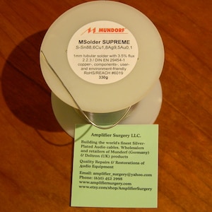 May include: A spool of silver-plated solder wire with a white label that reads "MSolder SUPREME S-Sn88,6Cu1,8Ag9,5Au0,1 1mm tubular solder with 3.5% flux 2.2.3/DIN EN 29454-1 copper, components, user- and environment-friendly ROHS/REACH #6019 330g". A green card with the text "Amplifier Surgery LLC. Building the world's finest Silver-Plated Audio cables. Wholesalers and retailers of Mundorf (Germany) & Deltron (UK) products Quality Repairs & Restorations of Audio Equipment Email: amplifier_surgery@yahoo.com Phone: (630) 452 2998 www.amplifiersurgery.com www.etsy.com/shop/AmplifierSurgery" is in front of the spool.