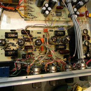 May include: Close-up of a vintage audio amplifier with multiple vacuum tubes, capacitors, resistors, and wires. The amplifier has a silver metal faceplate with four knobs. The circuit board is green and has a lot of wiring.