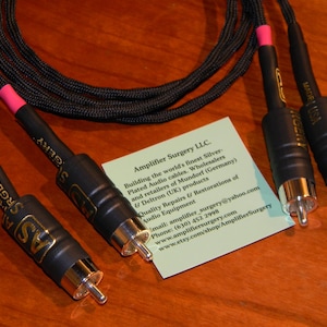 Puede incluir: Dos cables de audio RCA negros con conectores chapados en oro y aislamiento rosa. Los cables están etiquetados como "Amplifier Surgery" y "Made in USA".