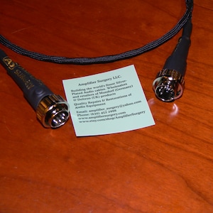 May include: A close-up of two black audio cables with silver-plated connectors. The cables are labeled "Amplifier Surgery LLC." and include contact information for the company.