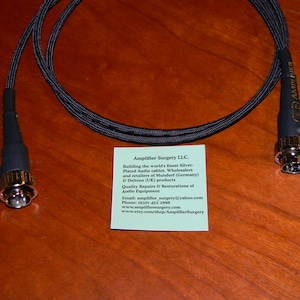May include: A black audio cable with gold-plated RCA connectors. The cable is coiled and has a black braided outer sheath. A business card for Amplifier Surgery LLC. is visible in the background.
