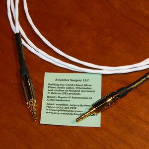 May include: Two white audio cables with gold-plated connectors. The cables are coiled and laid out on a wooden surface. A business card for Amplifier Surgery LLC. is visible in the foreground. The card includes the company's contact information and a description of their services.