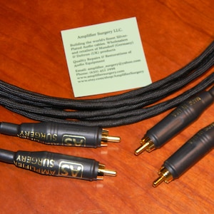Puede incluir: Tres cables de audio negros con conectores RCA dorados. Los cables están etiquetados como "Amplifier Surgery" y tienen una funda exterior trenzada negra.