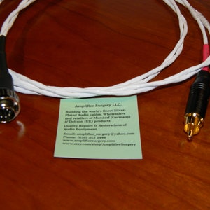 May include: White audio cable with a 5-pin DIN connector on one end and two RCA connectors on the other end. The cable is coiled and the connectors are gold-plated. A business card for Amplifier Surgery LLC. is visible in the background.