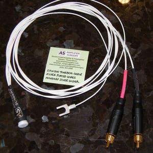 May include: A white tonearm cable with silver-plated wires and Mundorf silver solder. The cable is coiled and has two RCA connectors on the ends. The cable is labelled "Spin Din Tonearm Cable, Silver Plated Wires, Mundorf Silver Solder".