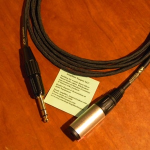 Puede incluir: Un cable de audio negro y plateado con un conector de 1/4 de pulgada en un extremo y un conector XLR en el otro. El cable está envuelto en tela trenzada negra. El cable está etiquetado "Amplifier Surgery LLC. Building the world's finest Silver Plated Audio cables. Wholesalers and retailers of Mundorf (Germany) &  Densen (UK) products. Quality Repairs & Restoration of Audio Equipment."