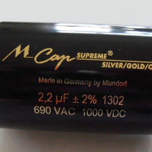 May include: A black cylindrical capacitor with gold lettering. The text reads "M-Cap SUPREME SILVER/GOLD/OIL Made in Germany by Mundorf 2,2 µF ± 2% 1302 690 VAC 1000 VDC".