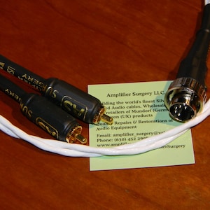 May include: Audio cables with black connectors and gold-colored tips, connected by white wires. The cables are labeled "SURGERY" and are on a wooden surface. A business card with text is also present.