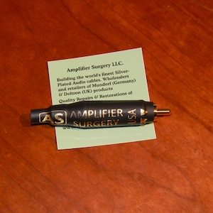 以下が含まれることがあります： 「AMPLIFIER SURGERY USA」と金色の文字が入った黒いオーディオケーブルコネクタ。オーディオケーブルと修理に関するテキストが書かれた緑色のカードの上にあります。背景は木製の表面です。