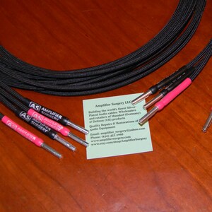 May include: Close-up of six black speaker cables with silver-plated connectors and pink heat shrink tubing. The cables are labeled "AS Amplifier Surgery".