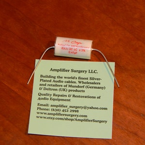 May include: A white capacitor with silver leads and the text "M Cap Audio MKP-Kondensator 630V DC AC ±3% 0.47µF" printed on it. A business card for Amplifier Surgery LLC. is in the background. The card includes the company's contact information and a description of their services, which include building silver-plated audio cables, wholesaling and retailing Mundorf and Deltron products, and providing quality repairs and restorations of audio equipment.