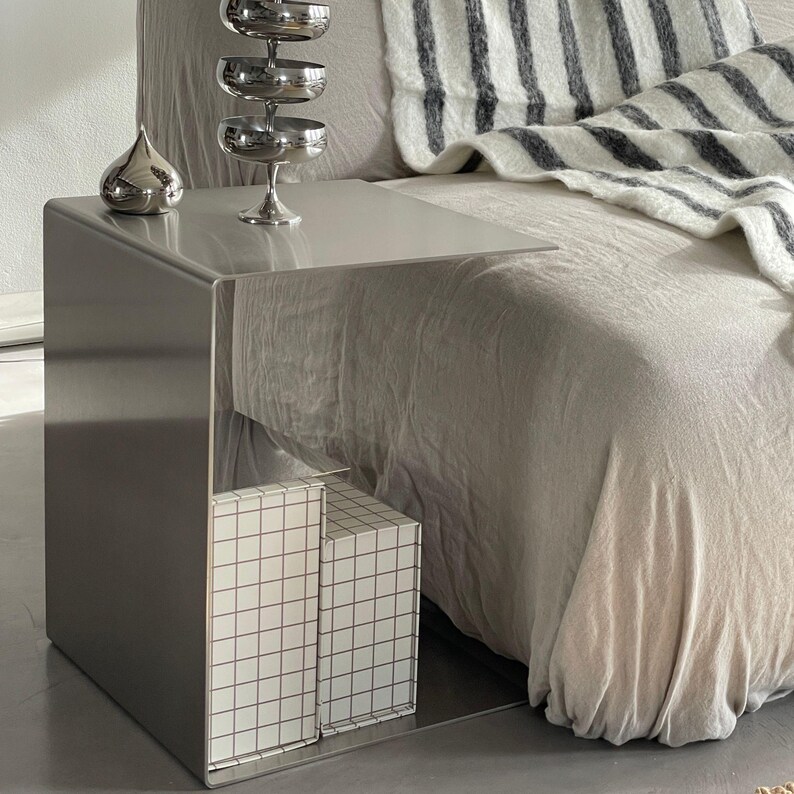 May include: A silver metal side table with a mirrored surface and two white boxes with a grid pattern on the bottom shelf. The table is next to a white and grey striped blanket on a bed.