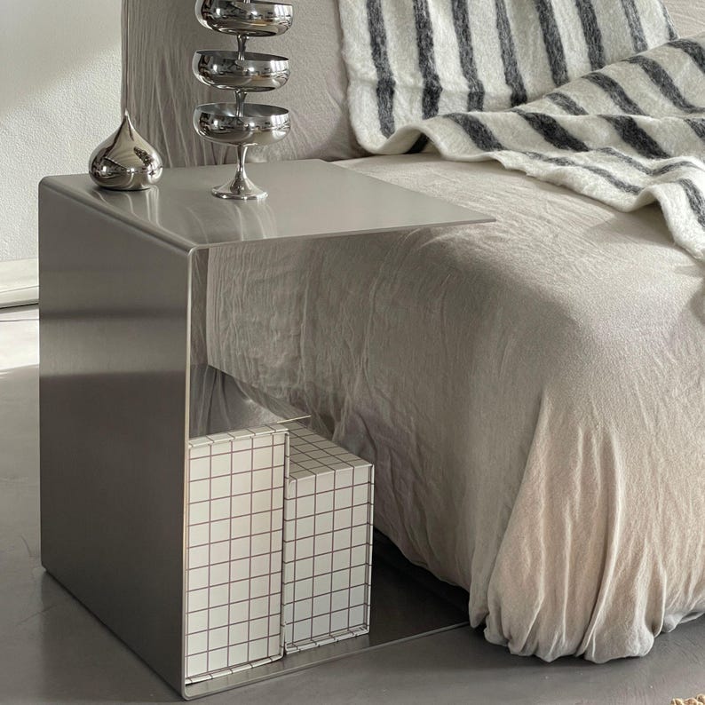 May include: A silver metal side table with a mirrored surface and two white boxes with a grid pattern on the bottom shelf. The table is next to a white and grey striped blanket on a bed.