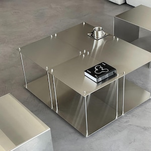 Peut inclure: Une table basse moderne en métal argenté, de forme carrée, avec une étagère inférieure. Un petit livre et une tasse en métal sur une soucoupe sont posés sur la table. La table est soutenue par de fines jambes en métal.