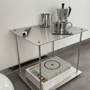 Op de afbeelding: Een stalen bijzettafel met twee niveaus met een zilveren koffiepot, een zilveren roomkan, een zilveren mok en een tube handcrème op het bovenste niveau. Het onderste niveau heeft een stapel tijdschriften en een boek met een cirkelvormig ontwerp op de omslag.