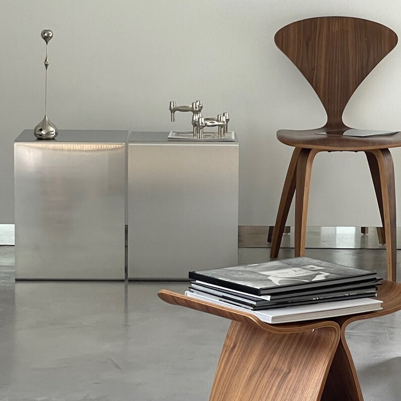 May include: Two silver metal side tables with a stack of books on a wooden stool in front of them. A silver metal sculpture sits on the table on the right. A wooden chair with a curved back is in the background.