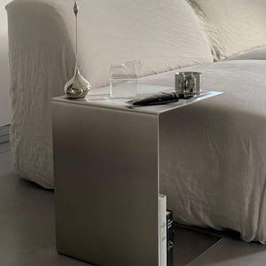 May include: A silver metal side table with a curved top and a book on the bottom shelf. The table is next to a white sofa and has a silver vase, a glass jug, and a silver cup on top.