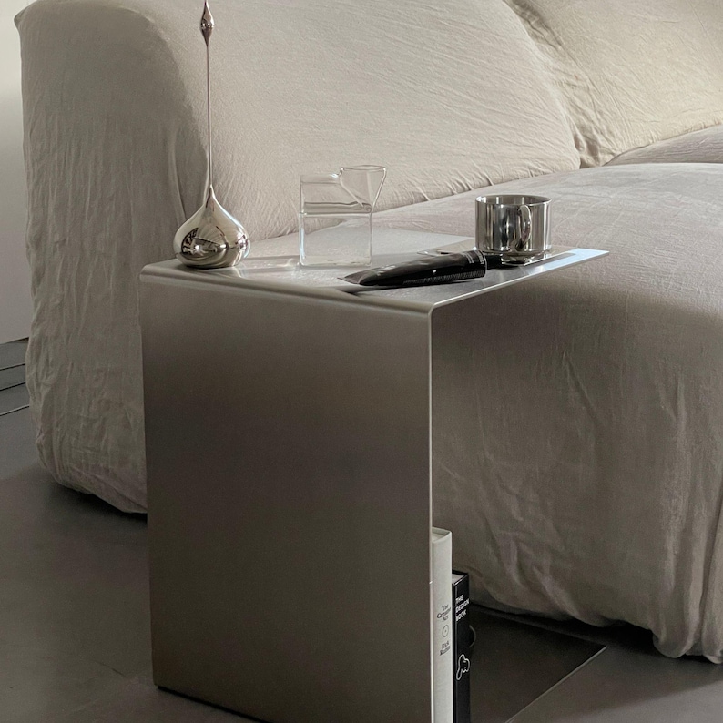 May include: A silver metal side table with a curved top and a book on the bottom shelf. The table is next to a white sofa and has a silver vase, a glass jug, and a silver cup on top.