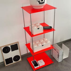 Op de afbeelding: Een viertraps rood rek met chromen steunen. De planken zijn gevuld met verschillende items, waaronder een witte doos met een lade, een zwart-witte telefoon en een witte doos met een lade gevuld met kleine witte blokken.