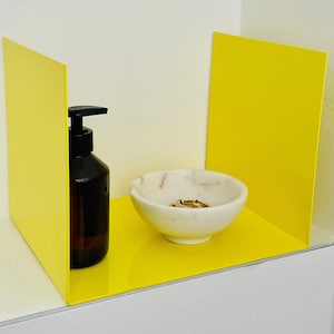 Könnte beinhalten: Ein gelbes Regal mit einer durchsichtigen Glasflasche mit Pumpspender, einer weißen Marmorschale mit goldenen Ringen und einem weißen Hintergrund.