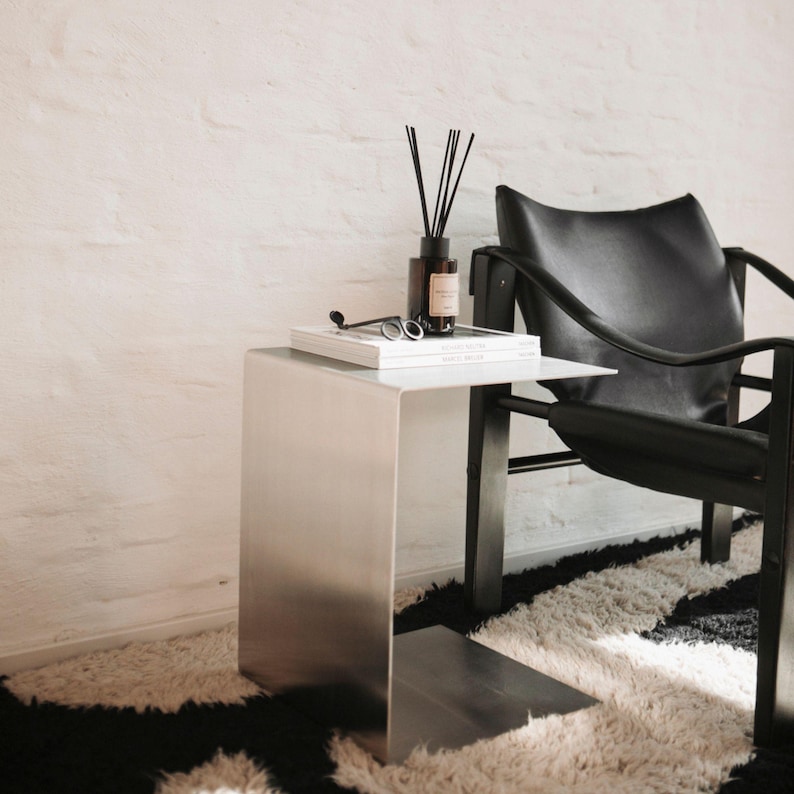 May include: A silver metal side table with a black leather chair and a white book on top. The table has a unique design with a shelf underneath. The chair is in the background and has a black leather seat and back.