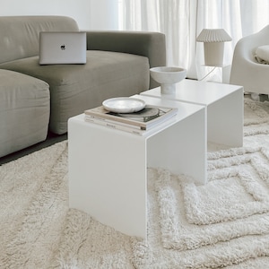 Op de afbeelding: Twee witte bijzettafels met een stapel boeken op de bovenkant van een tafel. De tafels staan op een wit, pluche tapijt voor een grijze bank. Een laptop staat op de bank.