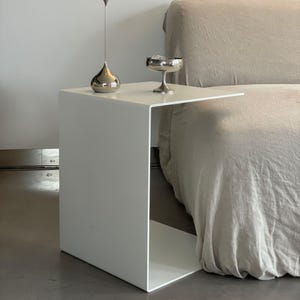 Könnte beinhalten: Ein weißer Metall-Beistelltisch mit einer rechteckigen Tischplatte und einem offenen Regal. Der Tisch steht neben einem beigen Sofa.