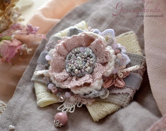 May include: A delicate pink and white flower brooch made with lace, fabric, and beads. The brooch is handmade and features a variety of textures and colours. It is a beautiful and unique piece of jewellery.