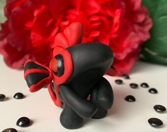 May include: A handcrafted black and red clay figurine with a stylized design. The figurine has a rounded body and head, with red accents on the eyes and wings. The background features a large red flower and scattered black beads.