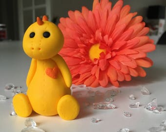 May include: A yellow clay figurine of a dragon with a heart on its chest, sitting on a white surface with clear glass shards and a large orange flower behind it.