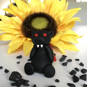 Peut inclure: Une figurine de chauve-souris noire avec des yeux rouges et des crocs blancs, portant une cape rouge. La figurine est posée sur une surface blanche avec des pierres noires.