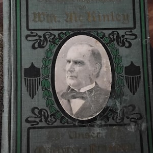 May include: Antique book with a teal cover featuring a portrait of a man in a suit and bow tie. The book's title, in gold lettering, reads "Die Lebensgeschichte von Wm. McKinley". Decorative green and black accents frame the portrait and text.