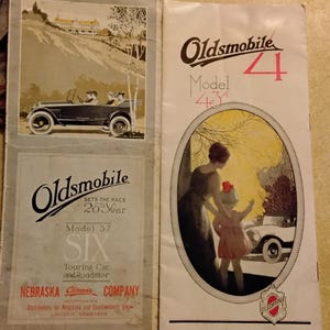 May include: Vintage Oldsmobile brochures. One features a black car on a road, the other shows a woman and child near a car. Text includes "Oldsmobile," "Model 37," and "Model 4."