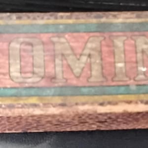 May include: A vintage wooden box with the word "DOMINO" in large, faded letters. The box has a red and green color scheme with star-shaped designs on each end. The edges of the box show signs of wear and tear.