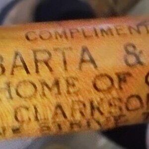 May include: A wooden rolling pin with the text "COMPLIMENTS OF BARATA & BARTA HOME OF GOOD MEATS CLARKSON, NEBR" etched into the wood. The rolling pin is a light brown color.