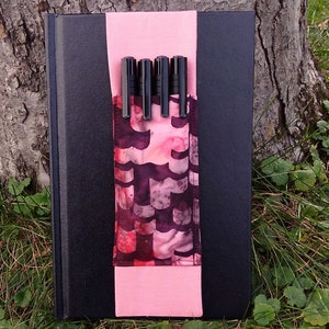 May include: A black notebook with a pink fabric pen holder with black pens inside. The notebook is sitting on a bed of green grass.