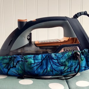 May include: A black, gray, and copper Shark iron rests inside a blue and green patterned iron cover. The iron is on a light blue surface with white polka dots. The iron cover has a drawstring closure.