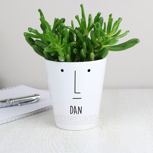 May include: White ceramic planter with a simple face design and the name "DAN" written below. The planter is filled with a green succulent plant.