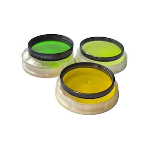 May include: Three circular camera lens filters with black metal frames, each resting on a clear plastic protective cap. The filters feature vibrant green, light yellow, and deep yellow glass elements, with the text 52-0.75 printed on the rims.