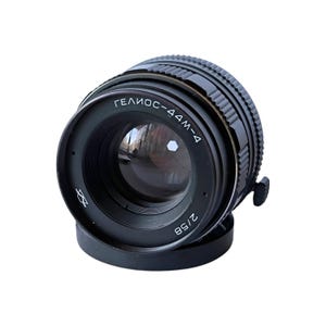 Peut inclure: Un objectif d'appareil photo Helios-44M-4 noir, avec une focale de 58 mm et une ouverture de f/2. L'objectif a une bague de mise au point striée et une base noire. Le texte "ГЕЛИОС-44М-4" et "2/58" est visible.