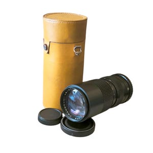 Può includere: Un obiettivo per fotocamera nero con lente argentata e due copriobiettivo neri. L'obiettivo è accanto a una custodia in pelle marrone chiaro. L'obiettivo ha il testo "ГРАНИТ-11".