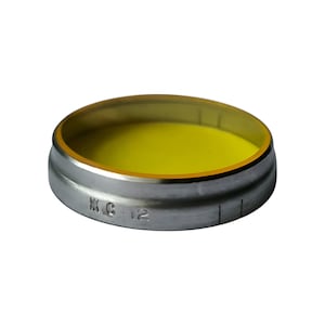 Op de afbeelding: Een rond, zilverkleurig metalen object met een gele binnenkant. Het object heeft een opstaande rand en een platte basis. De tekst "M.C. 12" is aan de zijkant gestempeld. De binnenkant is heldergeel.