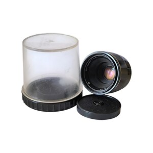 May include: A black camera lens with silver text, a black lens cap, and a clear plastic storage case. The lens is cylindrical with ridged edges. The lens cap is round with a textured surface.