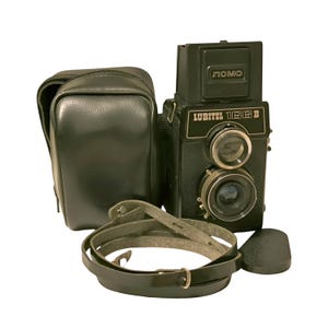 Peut inclure: Un appareil photo reflex à double objectif Lubitel 166B noir avec un étui et une sangle en cuir noir. L'appareil photo a un objectif argenté et une inscription argentée qui indique "LUBITEL 166B". L'appareil photo a également une plaque supérieure noire avec une inscription argentée qui indique "ЛОМО".
