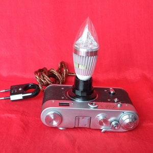 May include: A vintage silver camera with a black light bulb attached to the top. The light bulb has a clear glass cover and a silver base. The camera has a black cord with a plug attached.