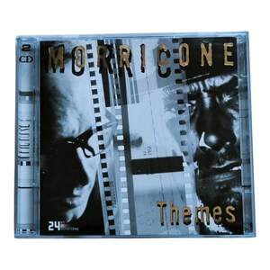 Puede incluir: Una caja de CD con el título "Morricone Themes" en letras doradas. La portada incluye imágenes en blanco y negro de un hombre y tiras de película. La caja es transparente, con una etiqueta "2 CD" y el texto "24 bit remastering".
